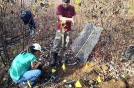 两名研究人员(1名男性站立), holding a shovel, 1 female squatting next to a whole) work in a prescribed burn site to plant potted red oak trees.